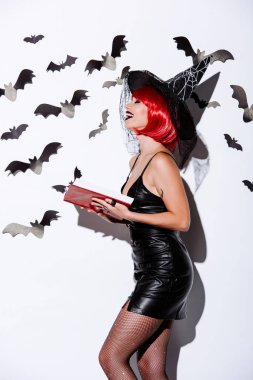 side view of laughing girl in black witch Halloween costume with red hair holding book near white wall with decorative bats clipart