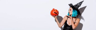 panoramic shot of sexy girl in black witch Halloween costume with blue hair blowing kiss to spooky carved pumpkin on white background clipart