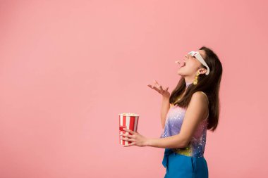 side view of happy young stylish disco girl in 3d glasses throwing popcorn in mouth isolated on pink clipart