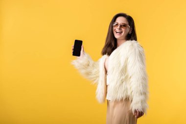 happy party girl in faux fur jacket and sunglasses holding smartphone with blank screen isolated on yellow clipart