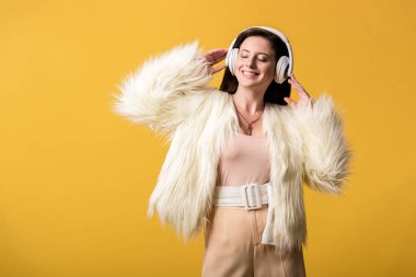 Sahte kürk ceketli, gülümseyen kız sarı kulaklıklı müzik dinliyor.