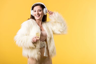 Sahte kürk ceketli gülümseyen kız kulaklıkla müzik dinliyor ve sarıda izole edilmiş bir bardak şampanya tutuyor.