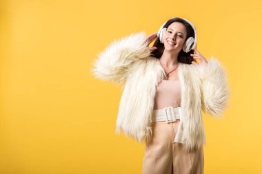Sahte kürk ceketli gülümseyen kız sarı renkteki kulaklıklarla müzik dinliyor.