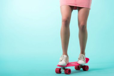 cropped view of girl in pink outfit standing on penny board on turquoise background clipart