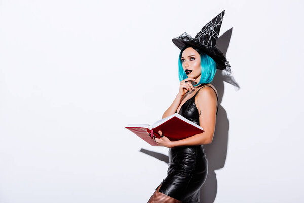 pensive sexy girl in black witch Halloween costume with blue hair holding book on white background