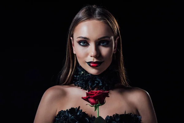 smiling scary vampire girl in black gothic dress holding red rose isolated on black