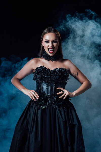 страшная девушка вампир с клыками в черно-готическом платье на черном фоне с дымом
