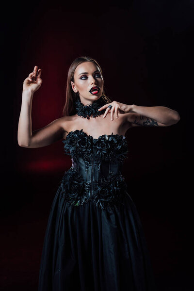 страшная девушка-вампир с клыками в черно-готическом платье на черном фоне
