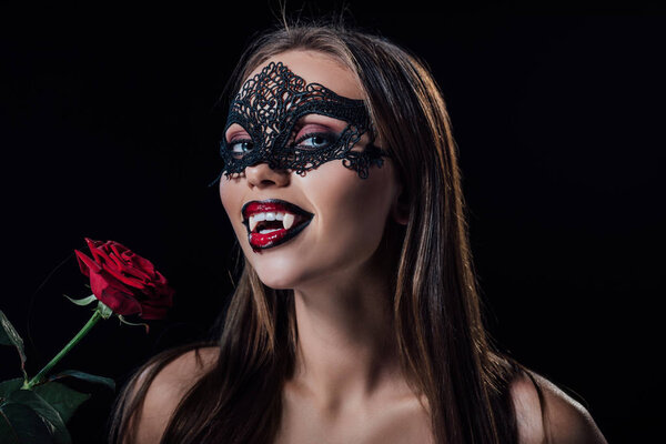 naked scary vampire girl in masquerade mask showing fangs and holding rose isolated on black