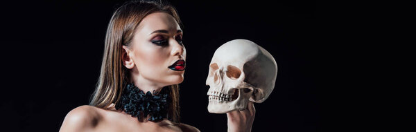 panoramic shot of scary vampire girl in black gothic dress looking at human skull isolated on black