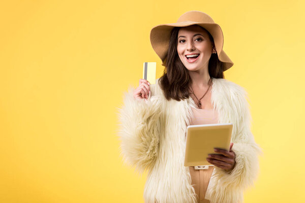 happy elegant girl in faux fur jacket and hat holding digital tablet and credit card isolated on yellow