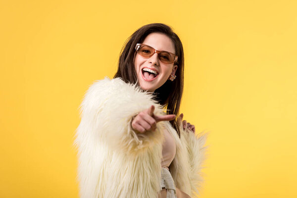 excited party girl in faux fur jacket and sunglasses pointing with finger at camera isolated on yellow