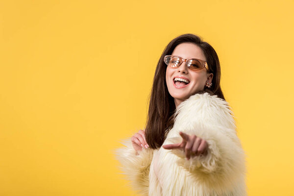 excited party girl in faux fur jacket and sunglasses pointing with finger at camera isolated on yellow