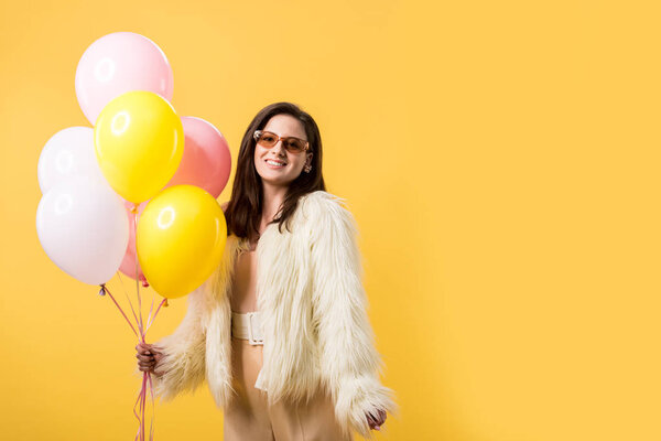 smiling party girl in faux fur jacket and sunglasses holding balloons isolated on yellow