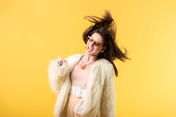 happy girl in faux fur jacket and sunglasses shaking head isolated on yellow