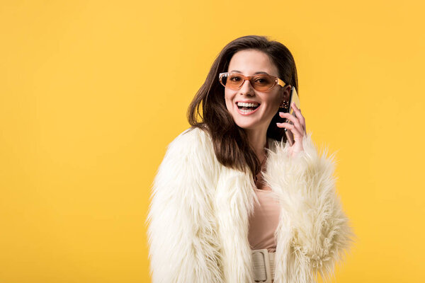 happy party girl in faux fur jacket and sunglasses talking on smartphone isolated on yellow