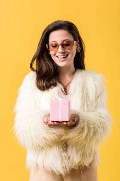 happy party girl in faux fur jacket and sunglasses looking at gift box isolated on yellow