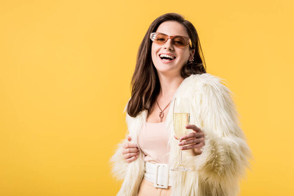 happy party girl in faux fur jacket and sunglasses with champagne isolated on yellow