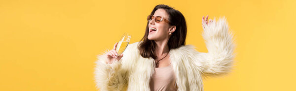 panoramic shot of happy party girl in faux fur jacket and sunglasses dancing with champagne isolated on yellow