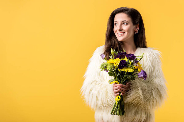 smiling girl in faux fur jacket holding bouquet of flowers isolated on yellow