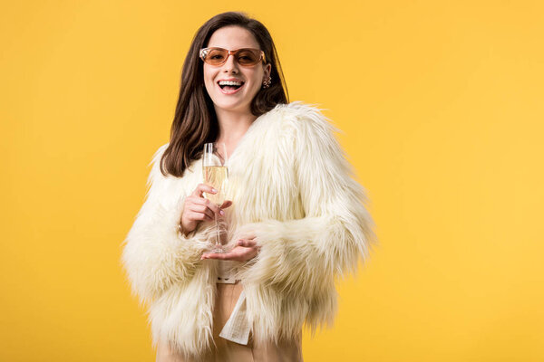 excited party girl in faux fur jacket and sunglasses with champagne isolated on yellow