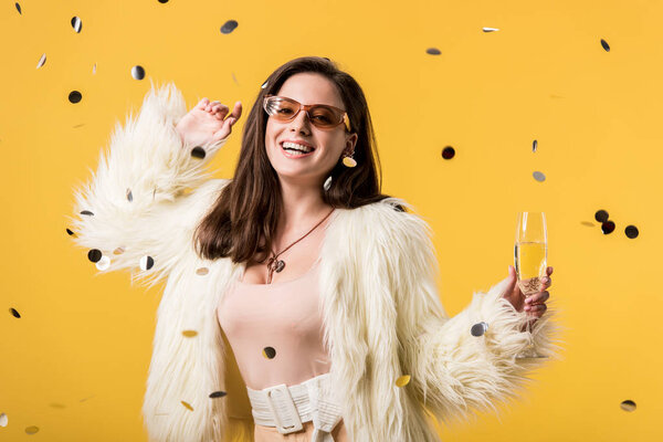 excited party girl in faux fur jacket and sunglasses standing under confetti with champagne isolated on yellow