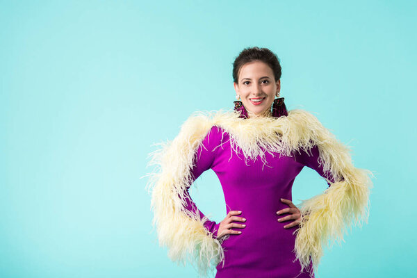 happy party girl in purple dress with feathers standing with hands on hips isolated on turquoise 