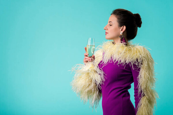 side view of party girl in purple dress with feathers holding glass of champagne isolated on turquoise 