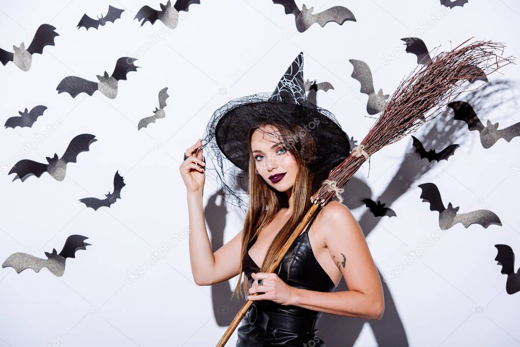 girl in black witch Halloween costume with broom near white wall with decorative bats