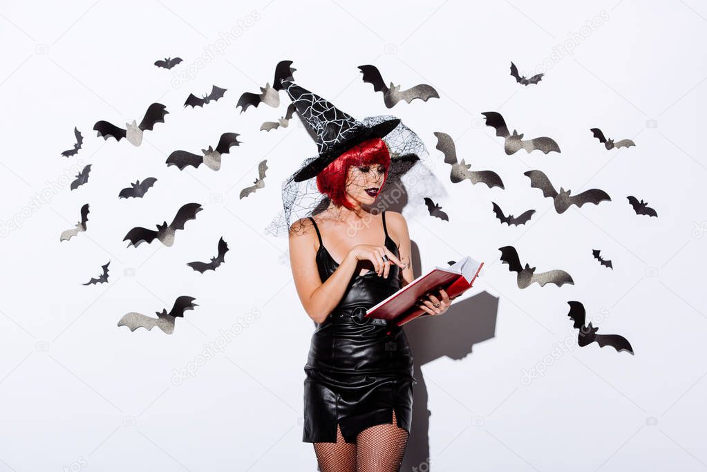 girl in black witch Halloween costume with red hair reading book near white wall with decorative bats