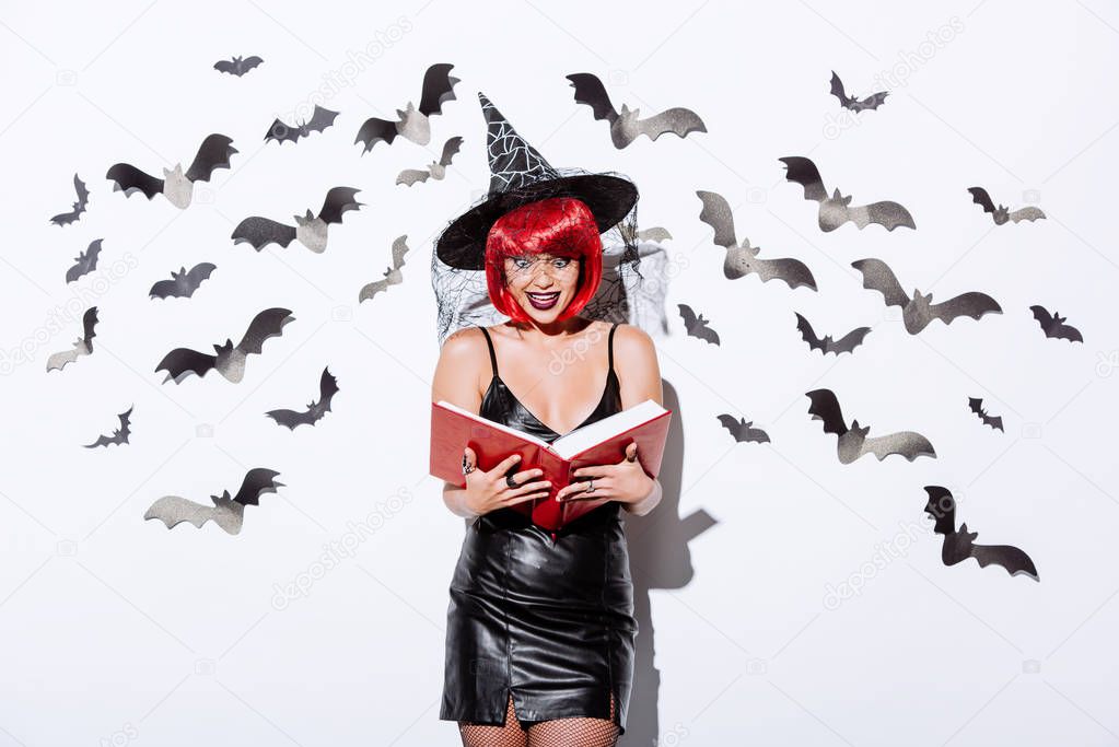 shocked girl in black witch Halloween costume with red hair holding book near white wall with decorative bats