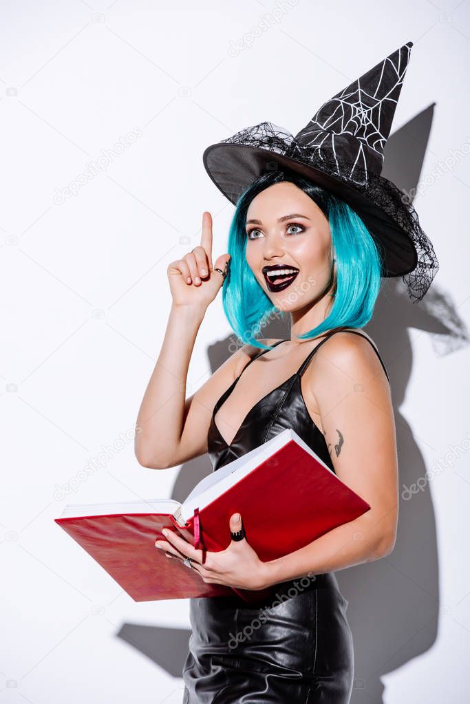 excited sexy girl in black witch Halloween costume with blue hair holding book and showing idea gesture on white background