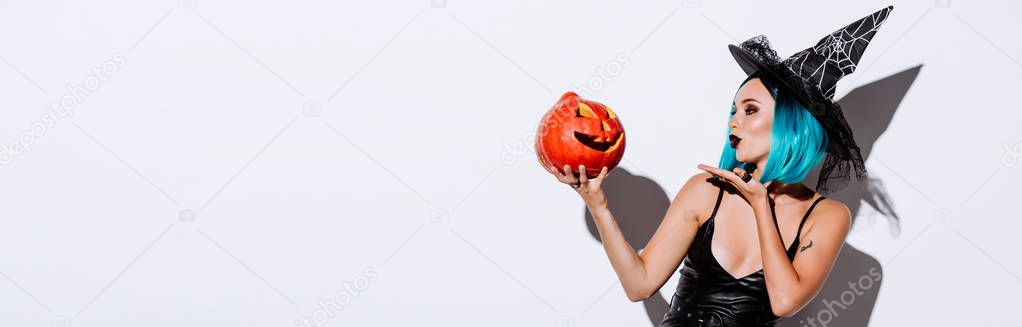 panoramic shot of sexy girl in black witch Halloween costume with blue hair blowing kiss to spooky carved pumpkin on white background