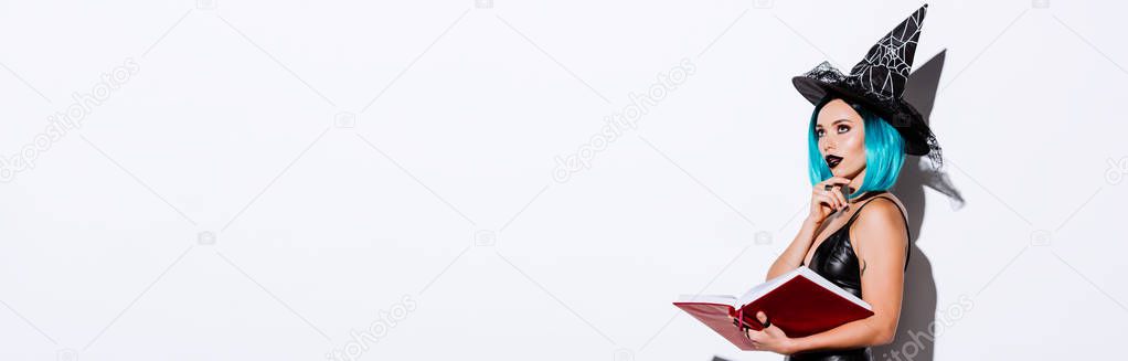 panoramic shot of pensive sexy girl in black witch Halloween costume with blue hair holding book on white background