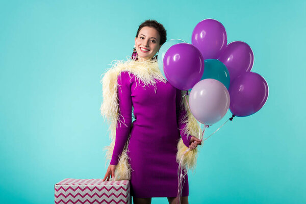 happy party girl in purple dress with feathers holding balloons near huge gift box isolated on turquoise 