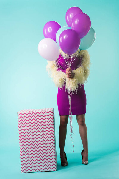 party girl in purple dress with feathers holding balloons in front of face near huge gift box on turquoise 