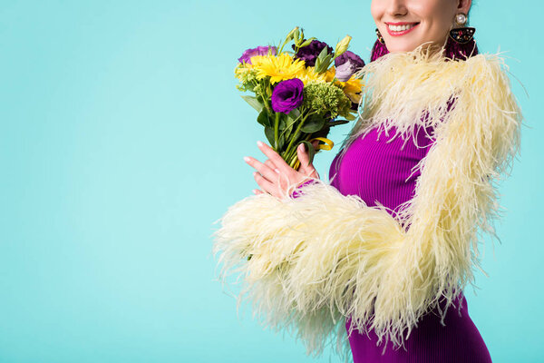 partial view of smiling party girl in purple dress with feathers holding bouquet of flowers isolated on turquoise 