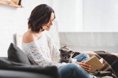 İskoç kedisiyle kanepede kitap okuyan çekici, gülümseyen bir kadın.  