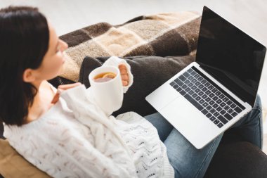 Elinde çay fincanı olan çekici bir kadın evinde dizüstü bilgisayardan film izliyor.