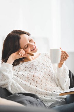 Kurabiye, kahve fincanı ve kitapla kanepede oturan mutlu kadın. 