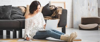 Kulaklıklı, güzel, gülümseyen bir kız. Evde dizüstü bilgisayar ve not defteriyle ders çalışıyor.