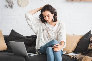 Çekici bir kız kanepede not defteri, kulaklık ve dizüstü bilgisayarla internetten düşünür ve çalışır.