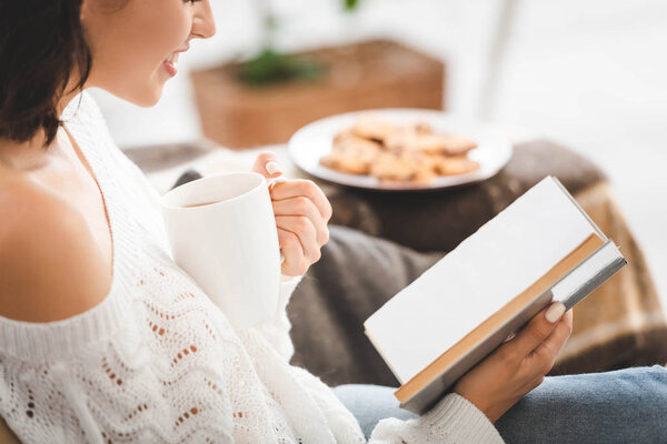 cropped view of girl reading book on sofa with cup of coffee and cookies