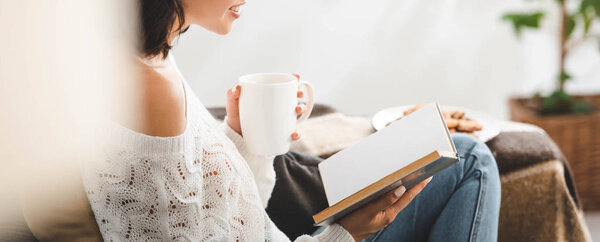 обрезанный вид девушки чтение книги на диване с чашкой чая и печенья
