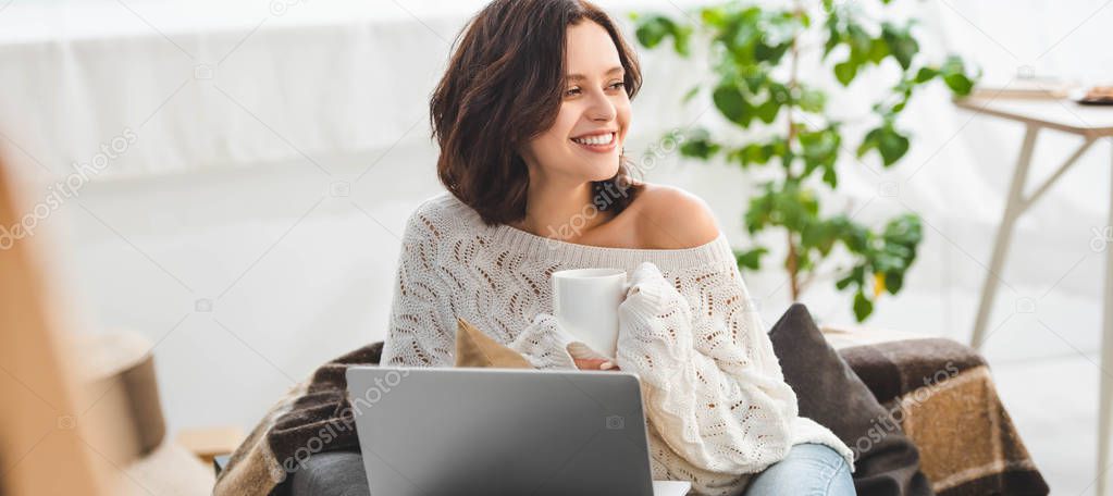 attractive smiling girl with cup of coffee using laptop at cozy home