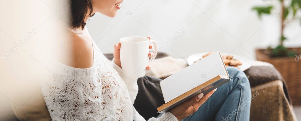 cropped view of girl reading book on sofa with cup of tea and cookies