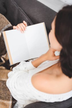 Evdeki kanepede battaniye giymiş kitap okuyan genç bir kadın.