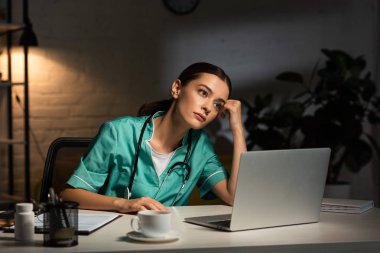 attractive and tired nurse in uniform sitting at table and looking away during night shift