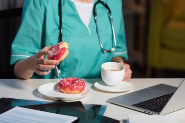 Gece vardiyasında üniformalı bir hemşirenin donut ve bardak tutarken çekilmiş görüntüsü.