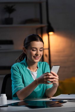Üniformalı gülümseyen hemşire masada oturuyor ve gece vardiyasında akıllı telefon kullanıyor.